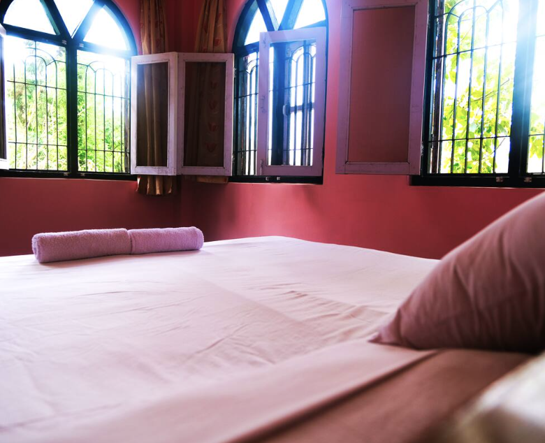 A maroon coloured room featuring arched windows and a comfortable bed. Out the windows you can see green trees and sunlight coming into the room.